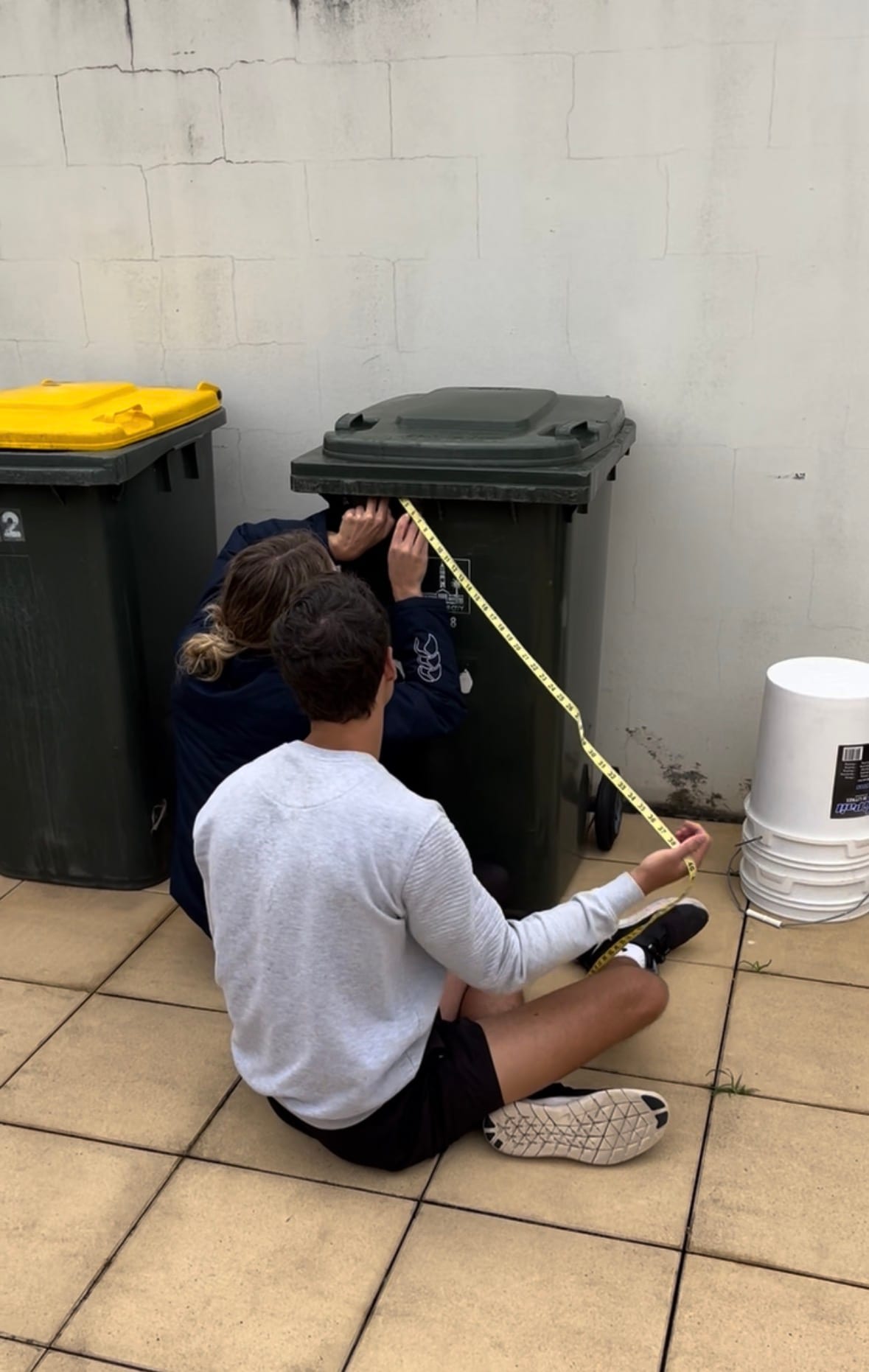 The founders crouched behind a green council bin, running a yellow tape measure over it